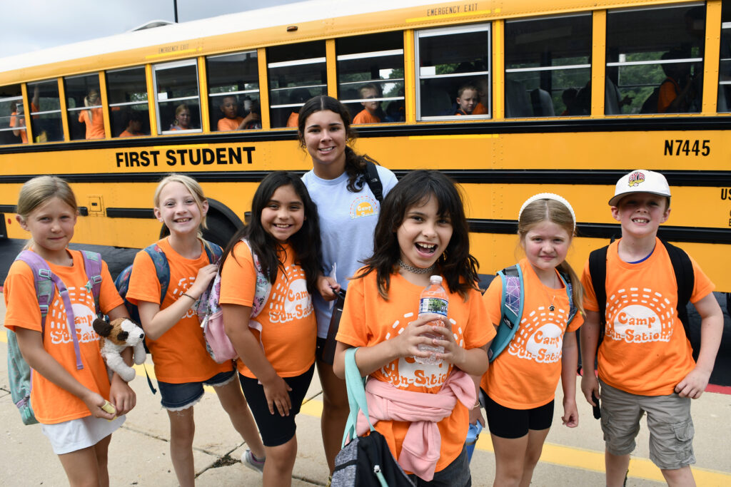 A group of summer campers and a counselor pose by the bus at the start of a field trip