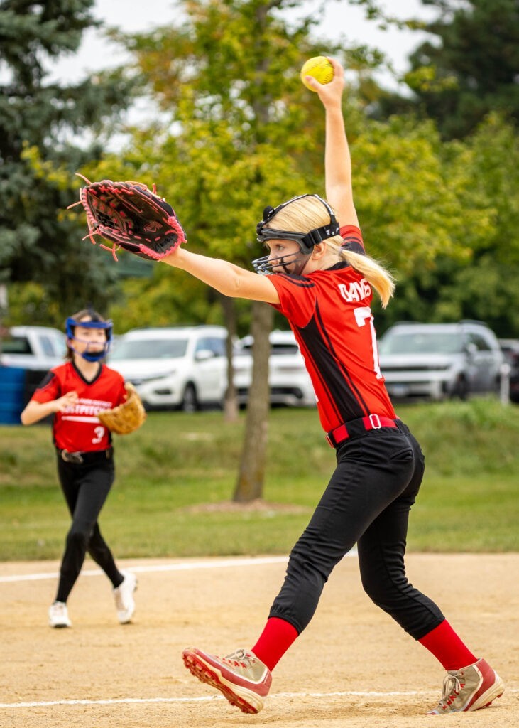 A softball pitcher winds up to throw a pitch with a teammate watching in the background