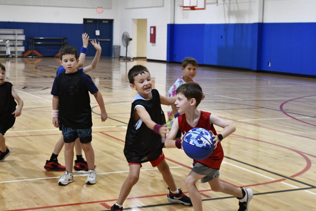 A young boy holds a basketball during a game in the gym while another boy tries to gain control of the ball as team members look on during summer Basketball Camp.