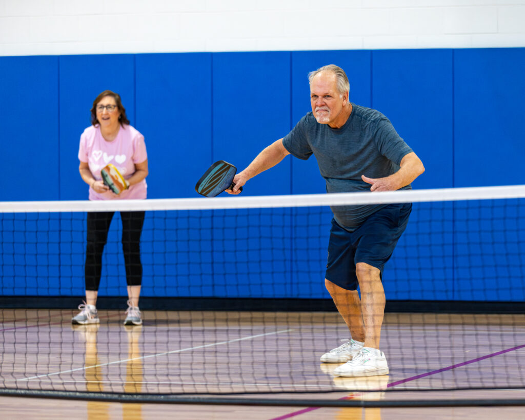 A senior male pickleball player leans toward the net, paddle in hand, his female partner watching, as he anticipates the path of the pickleball at Civic Center open gym