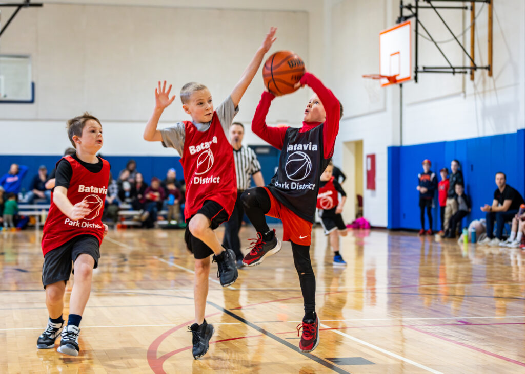 Three boys playing basketball in a youth league game, as one aims to make a basket and is blocked by a player of the opposing team