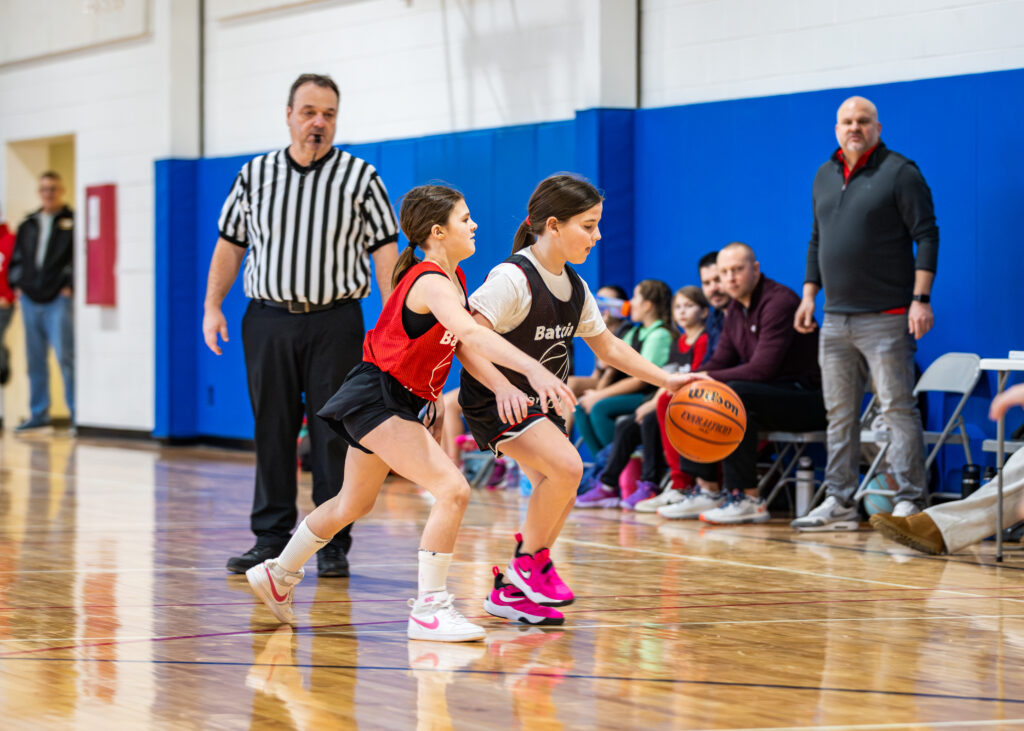 A girl on a youth basketball league team dribbles the ball as a player from the opposing team tries to gain control of the ball with the referee in the background